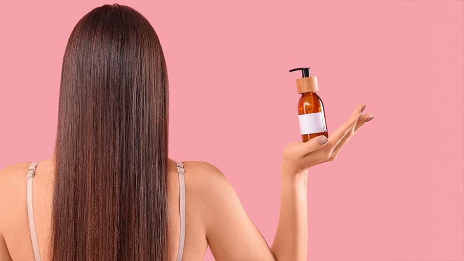 Kobieta stoi tyłem, ma proste włosy, w ręku trzyma butelkę z odżywką