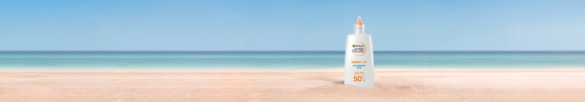 Biała butelka filtra Garnier Ambre Solaire na piaszczystej plaży, w tle ocean i niebo