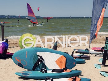 Deski surfingowe i SUP-y na słonecznej plaży, w tle windsurferzy na wodzie