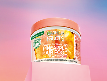 Biały słoik Garnier Fructis z pomarańczową pokrywką i motywem ananasa, na różowo-fioletowym gradiencie