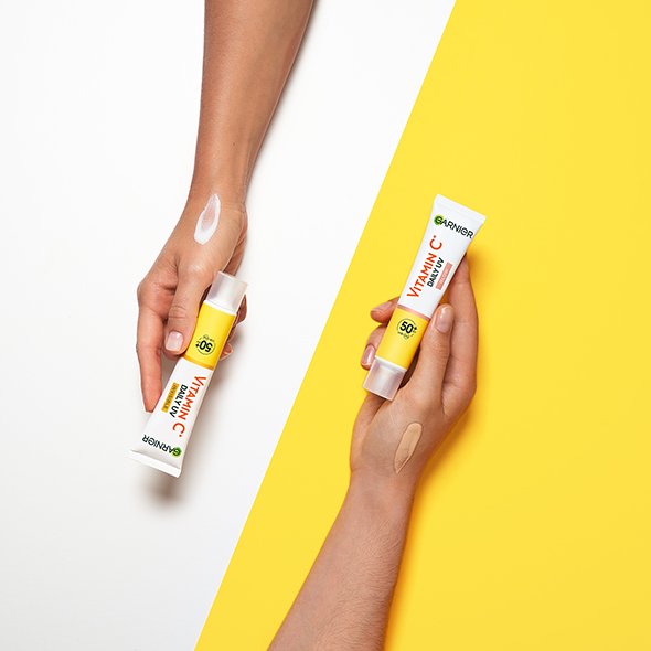 Dłonie trzymają tubki Garnier Vitamin C Daily UV SPF 50+ z białą i beżową próbką na przedramionach Dłonie trzymają tubki Garnier Vitamin C Daily UV SPF 50+ z białą i beżową próbką na przedramionach